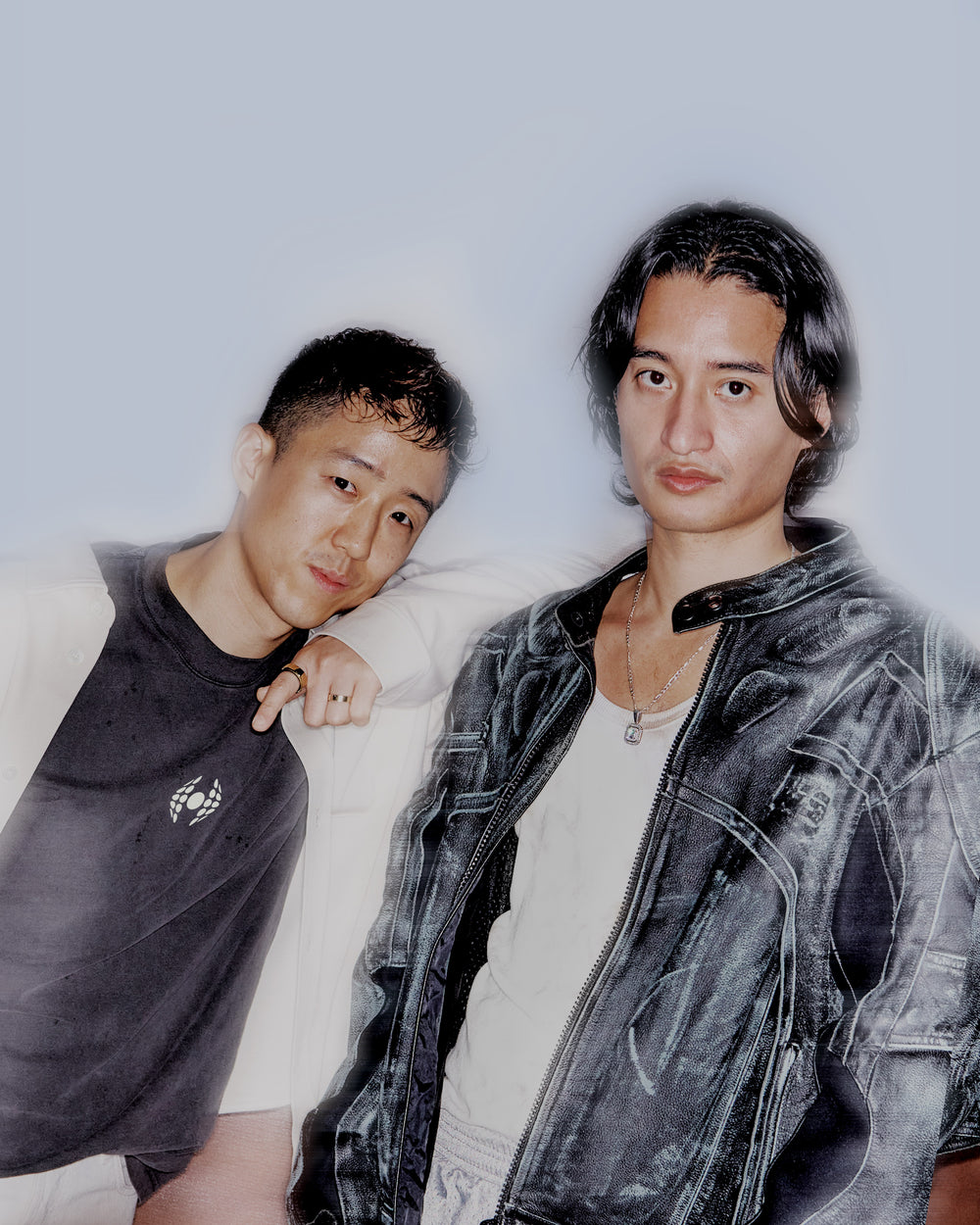 Two young men stand side by side against a light blue background; one wears a black T-shirt and cream jacket, the other wears a black leather jacket over a white shirt.